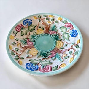 May include: A round, white ceramic plate with a raised floral design. The plate features a central teal-coloured well and a teal rim. The floral pattern includes pink, yellow, and blue flowers with green leaves and brown branches.