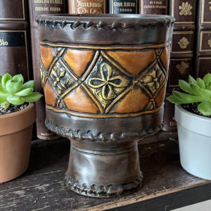 May include: A dark brown ceramic planter with a pedestal base, featuring a central band of raised, diamond-shaped patterns in a lighter tan colour with gold-toned floral accents and a stitched-look texture along the top and bottom rims.