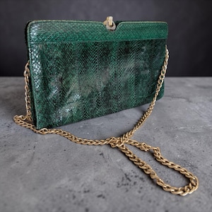 May include: A rectangular dark green clutch bag with a textured snakeskin pattern. The bag features a metallic gold-tone chain shoulder strap and a central top-mounted metal clasp closure. The exterior has a deep emerald green finish.