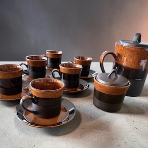 May include: A ceramic coffee set featuring a tall teapot, a lidded sugar bowl, and five matching cups with saucers. Each piece displays a two-tone design with a textured, speckled amber-brown upper section and a smooth, solid dark brown base.