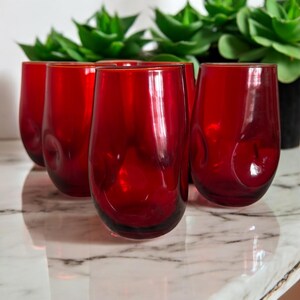 May include: A set of six stemless red wine glasses. The glasses are a deep red colour and have a rounded shape. The glasses are on a marble surface, with green plants in the background.