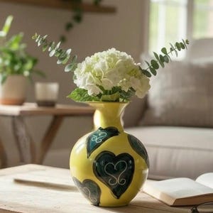 May include: A yellow ceramic vase with a round body and a narrow neck, decorated with dark teal heart and leaf designs. The vase holds a bouquet of white flowers and green foliage. The vase is on a wooden table.