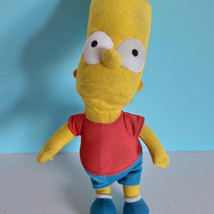 May include: A Bart Simpson soft toy. The toy has a yellow head and arms, a red top, blue shorts, and blue shoes. The toy has large white eyes with black pupils and a small nose.