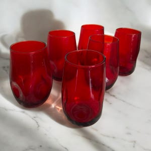 May include: A collection of six ruby-red stemless wine glasses. The glasses, made of glass, have a rounded design. They are displayed on a white marble surface. Each glass is approximately 10 cm tall.