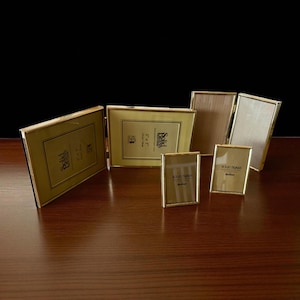 May include: A selection of gold-coloured brass picture frames in different sizes, displayed on a dark wooden surface. The rectangular frames are shown open and closed. Some frames have the text "Solid Brass".