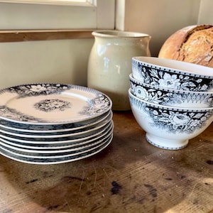 May include: A stack of six white ceramic plates and a stack of three white ceramic bowls, each featuring a black floral pattern along the rim and centre. The plates and bowls have a consistent decorative design with detailed botanical illustrations.