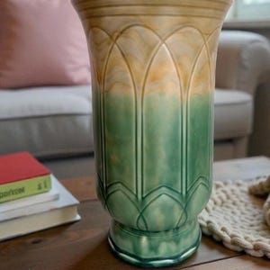 May include: A decorative vase with a flared top and a wide base. The vase has a green and tan colour scheme with a repeating arched pattern. The vase is on a wooden table.