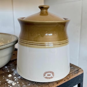 May include: A ceramic storage jar with a two-tone design featuring a glossy white base and a tan upper section with a matching lid. The jar has a circular stamp near the bottom that reads Bendigo Pottery 1858 Australia in brown text.