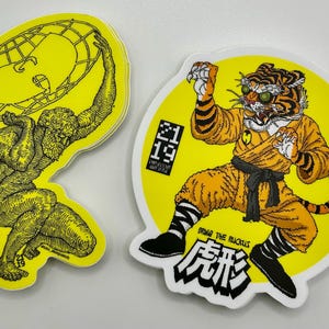 May include: Two stickers on a white surface. One is a yellow sticker with a black illustration of a figure holding a globe. The other is a yellow sticker with a tiger in a martial arts uniform. The tiger sticker has the text "BRING THE RUCKUS" and Japanese characters.