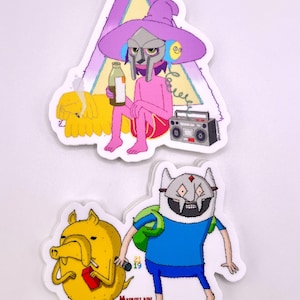 Puede incluir: Dos pegatinas con personajes de dibujos animados. Una muestra una figura con sombrero y máscara morados, sosteniendo una botella, con un perro amarillo y una radio. La otra representa dos personajes con el texto "MADVILLAIN TIME".