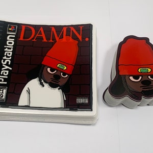 Puede incluir: Una pila de pegatinas con un personaje de dibujos animados que lleva una boina roja y una camisa blanca. La pegatina es una parodia de una portada de juego de Playstation y dice "DAMN." en la parte delantera.