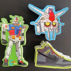 Peut inclure: Trois autocollants colorés illustrant un robot, une tête de robot et une chaussure. Le robot est blanc, rouge et bleu avec le texte "Gundam" sur sa jambe. La tête de robot est blanche, rouge et jaune. La chaussure est grise, noire et rouge avec le texte "DUNK" sur le côté.