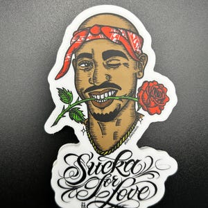 Op de afbeelding: Een sticker met een gestileerde illustratie van een man met een rode bandana en een rode roos in zijn mond. De tekst "Sucka for Love" staat eronder in een decoratief lettertype.