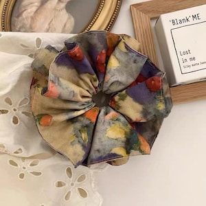 Könnte beinhalten: Ein großer Haargummi mit einem Aquarelldesign in Lila, Orange, Gelb und Grün. Der Haargummi ist aus einem weichen Stoff gefertigt und hat ein gerafftes, gerüschtes Aussehen. Der Text auf der Box lautet 'Blank Me' und 'Lost in me'.
