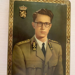 May include: A vintage tin box featuring a portrait of a man in military uniform. He is wearing glasses and has a serious expression. The box has a gold-coloured frame with a decorative design.