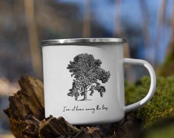 Può includere: Una tazza in smalto bianco con bordo e manico argentati, caratterizzata da un'illustrazione in bianco e nero di un grande albero e dal testo stampato "I am at home among the trees." in corsivo sulla parte anteriore.