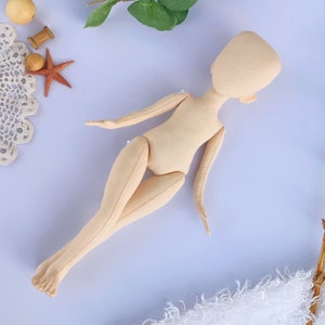 Peut inclure: Une poupée en tissu beige au design simple, avec une tête et des membres arrondis. La poupée est couchée sur une surface bleu clair.