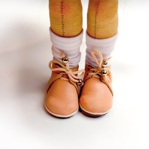 May include: Close-up of a pair of light peach-colored leather shoes with cream-colored soles and laces. The laces are tied in a bow and adorned with gold beads. The shoes are worn with white socks. The background is white.