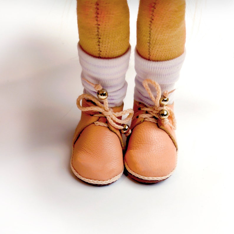 May include: Close-up of a pair of light peach-colored leather shoes with cream-colored soles and laces. The laces are tied in a bow and adorned with gold beads. The shoes are worn with white socks. The background is white.