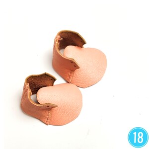 May include: Two pairs of baby shoes in a soft peach color. The shoes are made of leather, with a curved design and visible stitching. The interior of the shoes is a darker brown, and the shoes are set against a white background.