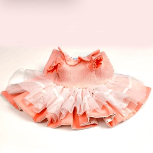 Puede incluir: Un vestido de color melocotón con una superposición de encaje blanco y un escote con volantes. El vestido tiene un patrón de puntos y es perfecto para una ocasión especial.