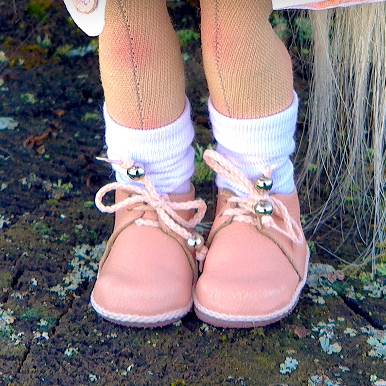 May include: A close-up of a doll's feet wearing pink leather shoes with white socks and a light pink bow. The shoes have silver-colored accents and are tied with a light pink string. The doll's legs are covered in tan-colored fabric.