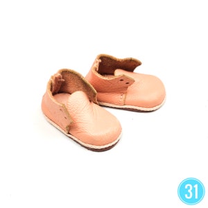 May include: A pair of peach-colored baby shoes made of soft leather. The shoes have a rounded toe, a stitched sole, and a folded-over collar. The shoes are designed for infants and are set against a white background.
