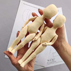Könnte beinhalten: Zwei Puppenkörper aus beigem Stoff mit beweglichen Gliedmaßen werden in den Händen einer Person gehalten. Die Puppen sind über einem bedruckten Schnittmusterbogen positioniert. Die Puppen befinden sich in den frühen Stadien der Konstruktion, mit sichtbaren Nähten und einem weichen, unfertigen Aussehen.