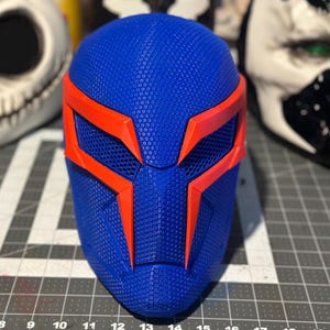 May include: A blue and orange superhero mask with a textured surface. The mask features a geometric design with orange accents around the eyes and forehead. Other masks are visible in the background.
