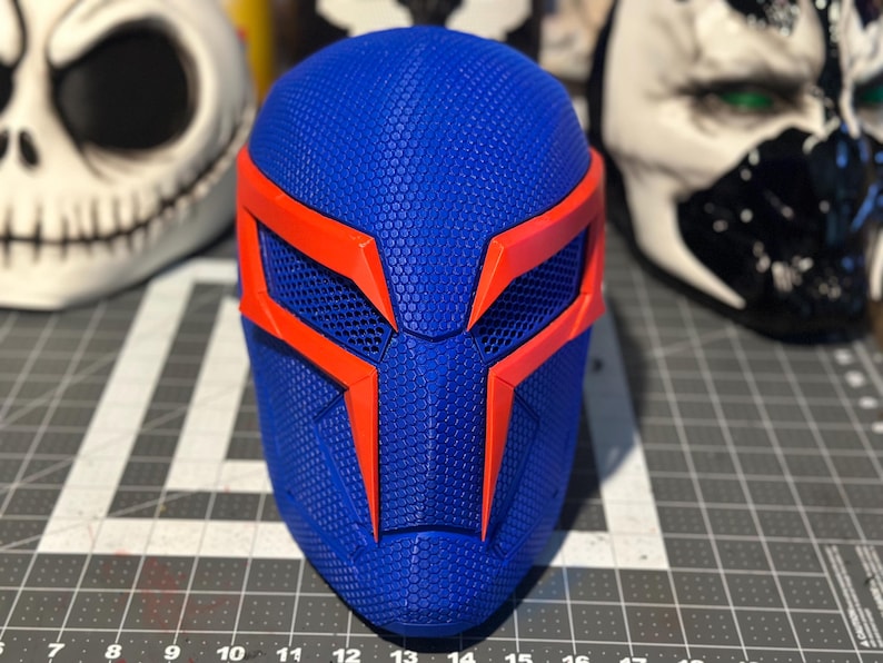 May include: A blue and orange superhero mask with a textured surface. The mask features a geometric design with orange accents around the eyes and forehead. Other masks are visible in the background.
