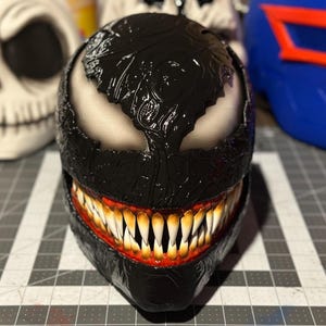 May include: A black and white Venom mask with a detailed textured surface and a red and white mouth with sharp teeth.