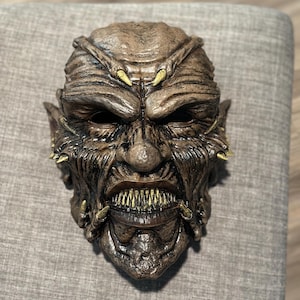 May include: A detailed brown monster mask with sharp teeth and yellow accents. The mask has a textured surface and is designed with a menacing expression, suitable for Halloween or costume parties.
