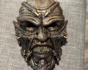 Pode incluir: Uma máscara de monstro castanha detalhada com dentes afiados e detalhes amarelos. A máscara tem uma superfície texturizada e é projetada com uma expressão ameaçadora, perfeita para o Halloween ou festas à fantasia.