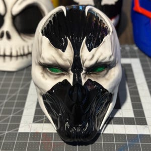 May include: A black and white mask with green eyes. The mask has a detailed design with a black and white pattern.