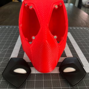 May include: A red 3D printed Deadpool mask with two black eye pieces. The eye pieces have white eye holes.