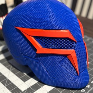 May include: A blue and orange helmet with a textured surface. The helmet features a geometric design with orange accents around the eye area and a mesh vent. Likely a cosplay or costume piece.