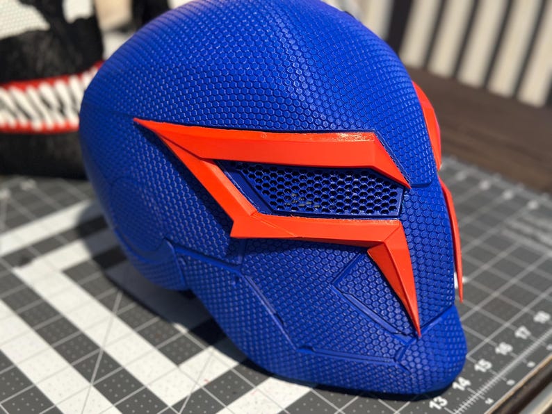 May include: A blue and orange helmet with a textured surface. The helmet features a geometric design with orange accents around the eye area and a mesh vent. Likely a cosplay or costume piece.