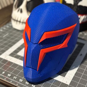 May include: A blue and orange superhero mask with a textured surface. The mask has a geometric design with orange accents around the eyes and edges. The mask is likely for cosplay or costume use.
