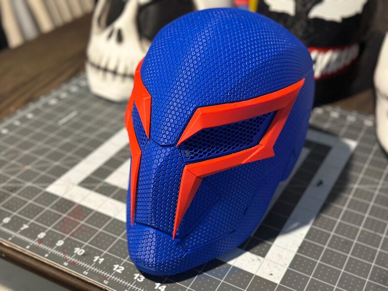 May include: A blue and orange superhero mask with a textured surface. The mask has a geometric design with orange accents around the eyes and edges. The mask is likely for cosplay or costume use.
