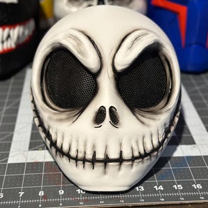 May include: A white plastic Halloween mask with a black mesh covering the eyes. The mask is sculpted to look like a skull with a wide grin and a stitched-up mouth.