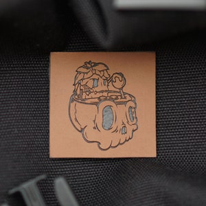 May include: A brown leather patch with a black and white illustration of a skull with a leafy plant growing out of the top.