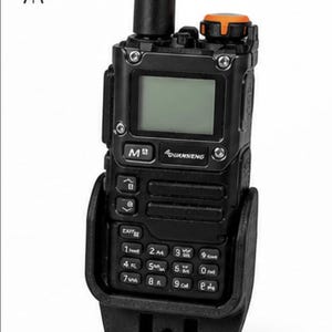 Puede incluir: Radio bidireccional portátil negra con antena negra y funda negra. La radio tiene una pequeña pantalla y un teclado con números y símbolos. La parte superior de la radio tiene un dial naranja. El texto "SIGNAL MODS" es visible.