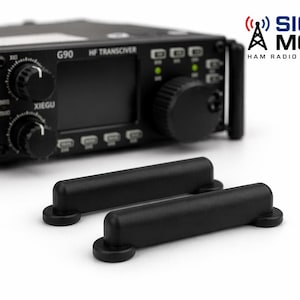 Może przedstawiać: Czarny transceiver HF XIEGU G90 z dwiema czarnymi stopkami magnetycznymi. Transceiver ma wiele pokręteł i przycisków. Logo Signal Mods znajduje się w prawym górnym rogu.