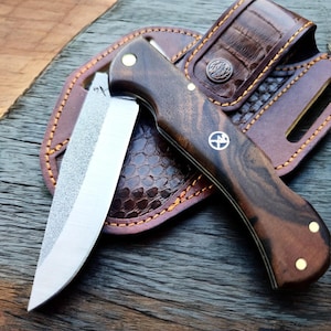 Könnte beinhalten: Ein Klappmesser mit einem dunklen Holzgriff und einer silbernen Klinge, das auf einer braunen Lederscheide liegt. Der Griff hat Messingnieten und ein dekoratives Emblem. Die Scheide hat ein strukturiertes Muster und genähte Kanten.