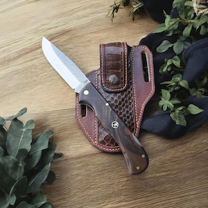 Op de afbeelding: Een vouwmes met een houten handvat en een roestvrijstalen lemmet, gepresenteerd met een bruine leren schede. Het handvat heeft messing klinknagels en een decoratief embleem. De schede heeft een textuurpatroon en een riemlus.