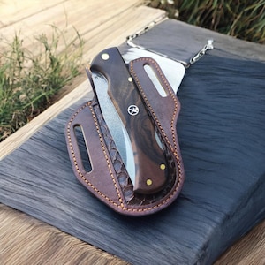 Könnte beinhalten: Ein Klappmesser mit dunklem Holzgriff und silberner Klinge, eingebettet in einer braunen Lederscheide. Die Scheide hat dekorative Nähte und ein strukturiertes Muster. Das Messer ist für den täglichen Gebrauch konzipiert.
