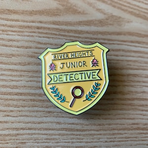Pode incluir: Um alfinete de esmalte amarelo e preto em forma de escudo. O alfinete diz "River Heights Junior Detective" com uma lupa e folhas de louro. O alfinete está em uma superfície de madeira.