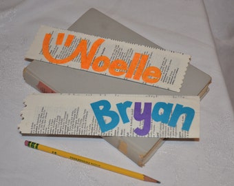 Puede incluir: Dos marcapáginas personalizados con los nombres "Noelle" y "Bryan" escritos en letras de colores sobre un fondo blanco. Los marcapáginas están hechos de páginas de diccionario.