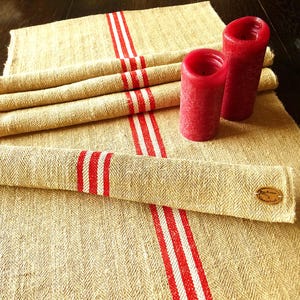 Könnte beinhalten: Beige Leinentischläufer mit drei vertikalen rot-weißen Streifen. Zwei rote Säulenkerzen sind auf dem Läufer platziert. Der Läufer ist gefaltet und aufgerollt, mit einem kleinen Holzanhänger versehen.