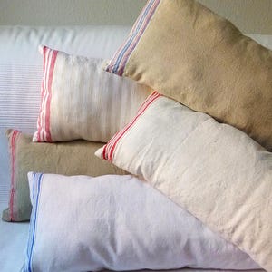 Pode incluir: Uma coleção de almofadas retangulares em vários tons de bege, branco sujo e creme. Cada almofada apresenta uma textura tecida distinta e é acentuada com listras verticais em vermelho, azul ou uma combinação de vermelho e azul numa borda.